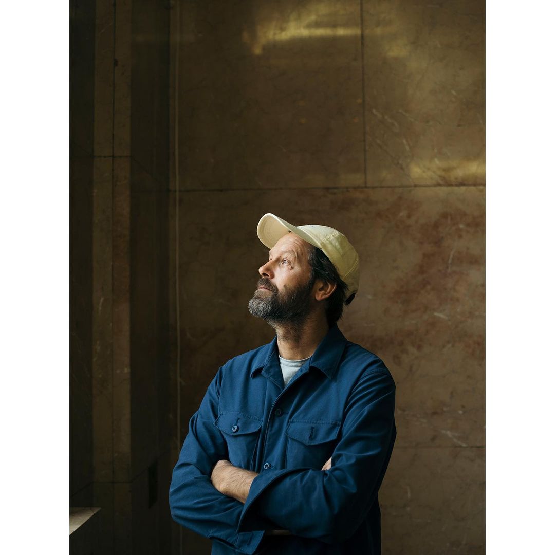 Man in a baseball cap and blue button-down shirt poses indoors with arms crossed, looking up, with a marble-like wall in the background.