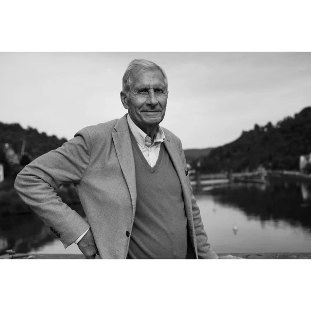 Elderly man in jacket and sweater poses outdoors on bridge with water and green hills in background. Black and white portrait photo.