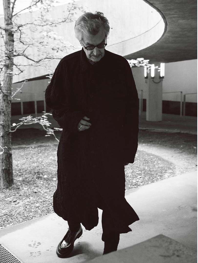 Elderly man in a long, dark coat walks down concrete steps next to a tree in a monochrome, architectural setting.