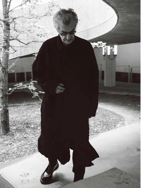 Elderly man in a long, dark coat walks down concrete steps next to a tree in a monochrome, architectural setting.