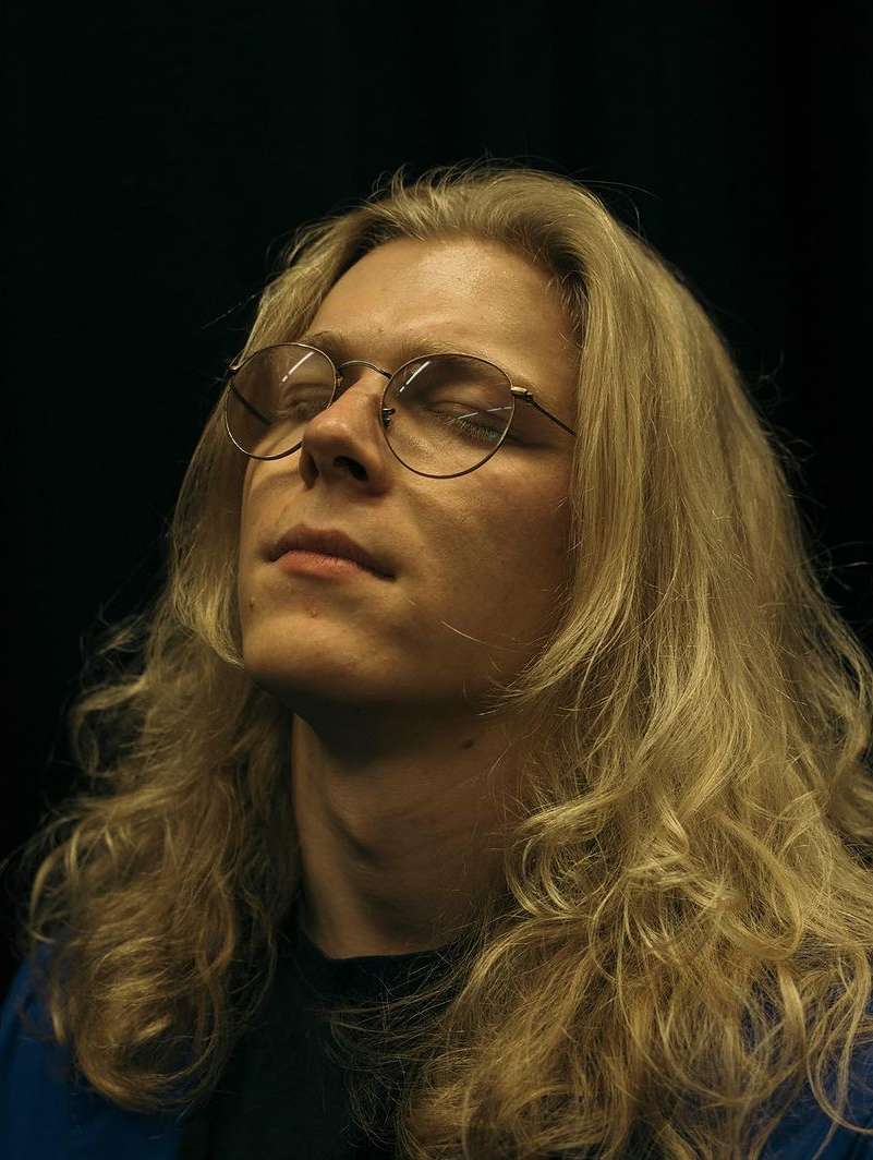 Portrait of adult male with blond, shoulder-length hair and glasses, eyes closed, against a dark background in indoor lighting.
