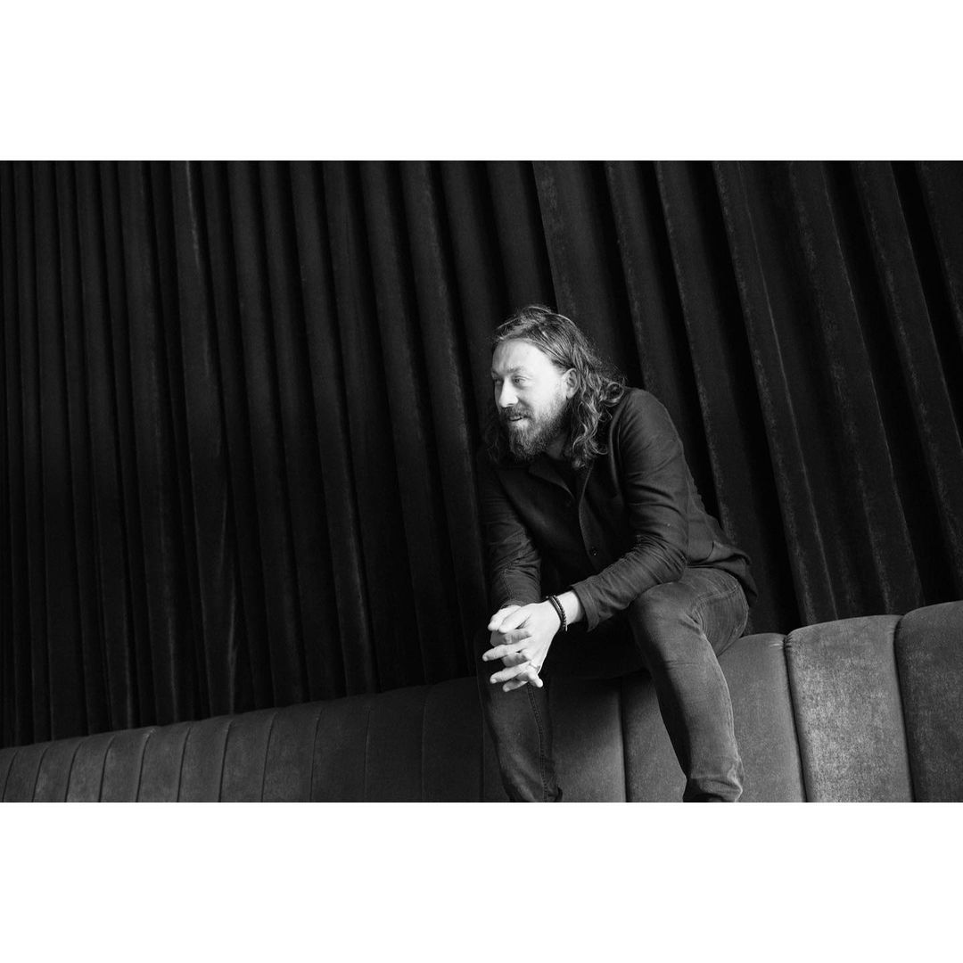 Monochrome portrait of a seated adult male with long hair and a beard, wearing dark clothing against a black curtain backdrop.