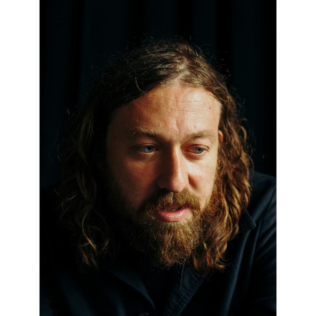 Close-up portrait of an adult man with a beard and long hair, wearing a dark shirt, against a dark background.