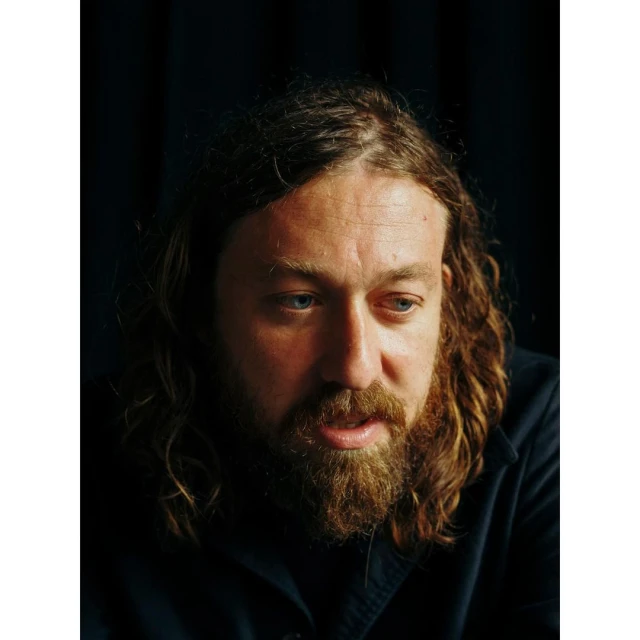 Close-up portrait of an adult man with a beard and long hair, wearing a dark shirt, against a dark background.