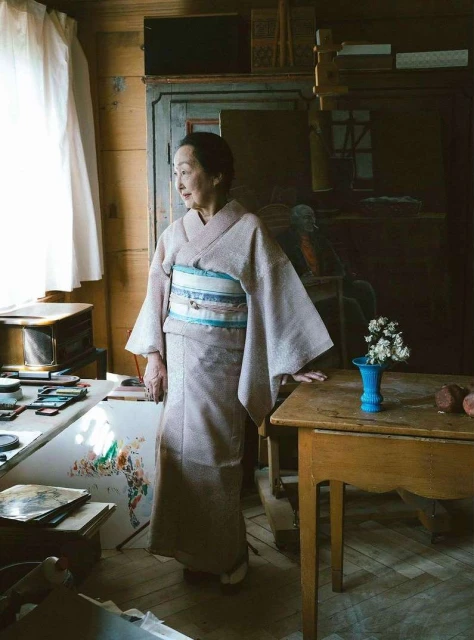 Elderly Asian woman in a light pink kimono standing in a rustic room near a window, with a vase of white flowers on a table.
