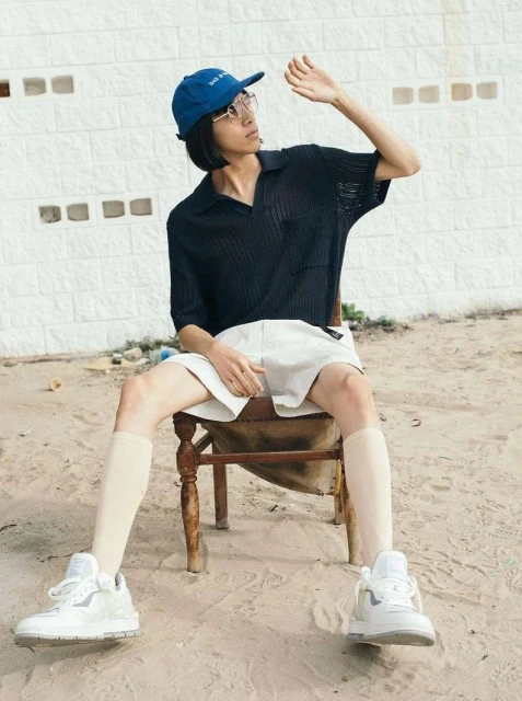 A young adult male sits on a wooden chair outdoors, wearing a blue cap, glasses, navy polo shirt, shorts, beige socks and white sneakers.