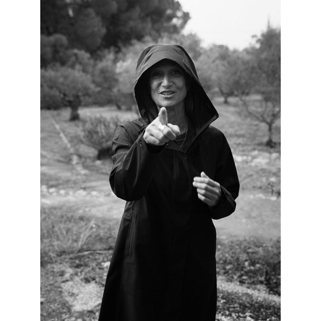 Woman in hooded black raincoat points at the camera in a monochrome, outdoor portrait set against olive trees in a field.