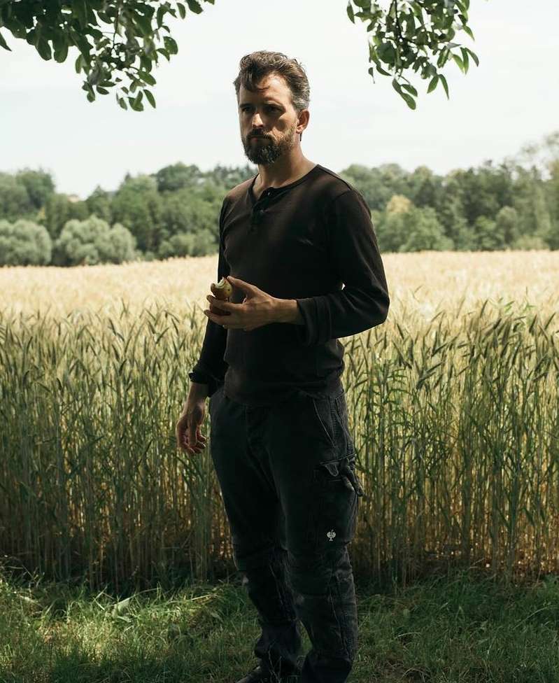 A man with a beard stands in a wheat field holding an apple, wearing a black shirt and pants. Outdoors, full shot, daytime.