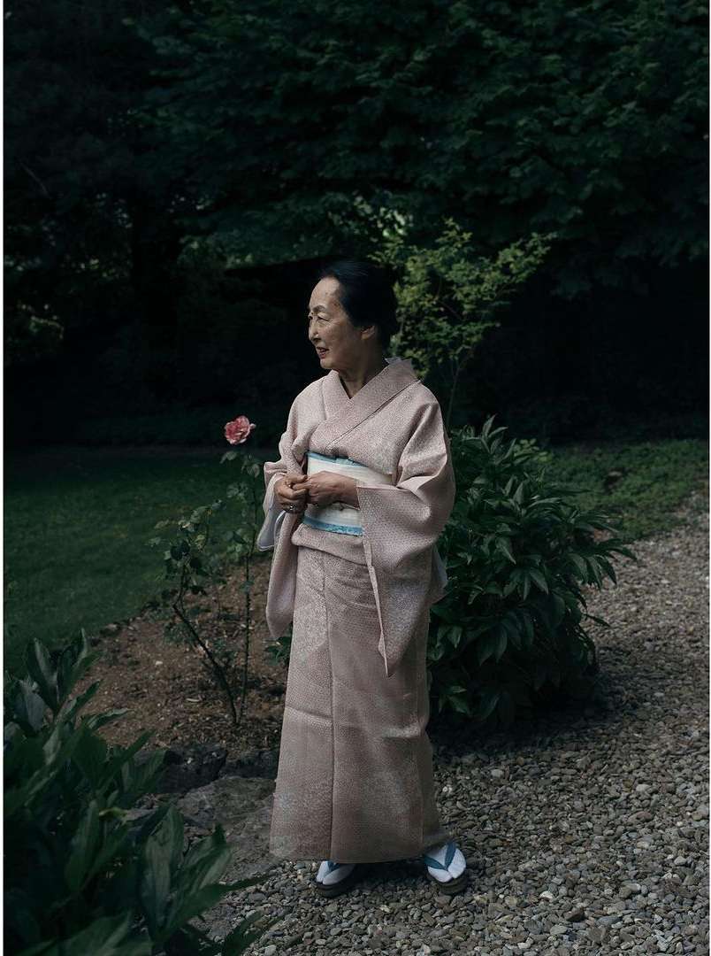 Elderly woman in a light pink kimono stands in a lush garden with greenery and a gravel path in a serene outdoor setting.