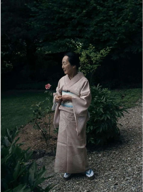 Elderly woman in a light pink kimono stands in a lush garden with greenery and a gravel path in a serene outdoor setting.
