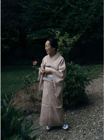 Elderly woman in a light pink kimono stands in a lush garden with greenery and a gravel path in a serene outdoor setting.