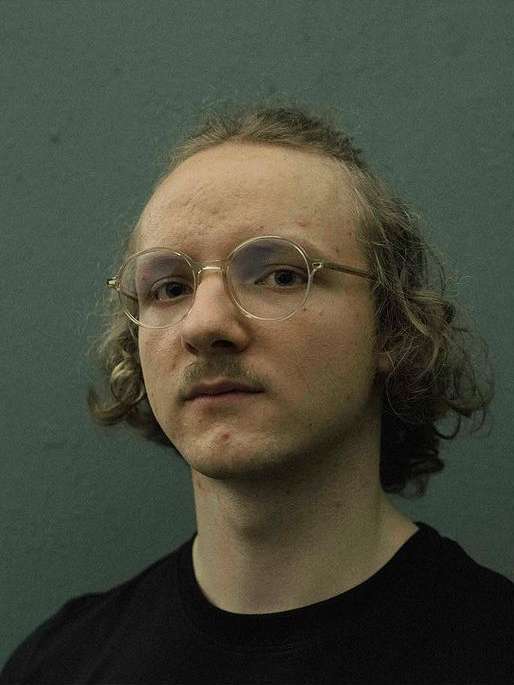 Portrait of a man wearing round wire-rimmed glasses and a black t-shirt against a dark textured wall.
