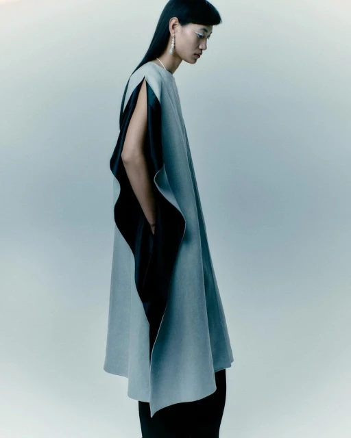 Fashion shot of an Asian female model in studio wearing a light grey cape with black lining, long black hair, and silver earrings.