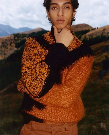 Young man wearing a textured orange and black patterned sweater posing outdoors against rolling green hills and cloudy sky.
