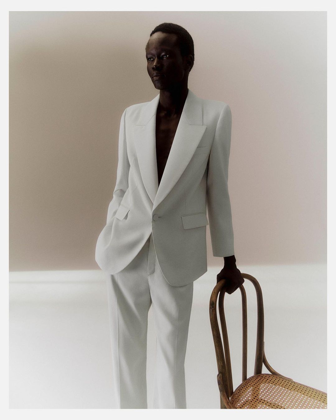 Black man wearing a white suit jacket and trousers resting his hand on a wooden chair in Paris by Matthieu Delbreuve
