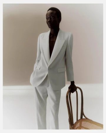 Black man wearing a white suit jacket and trousers resting his hand on a wooden chair in Paris by Matthieu Delbreuve