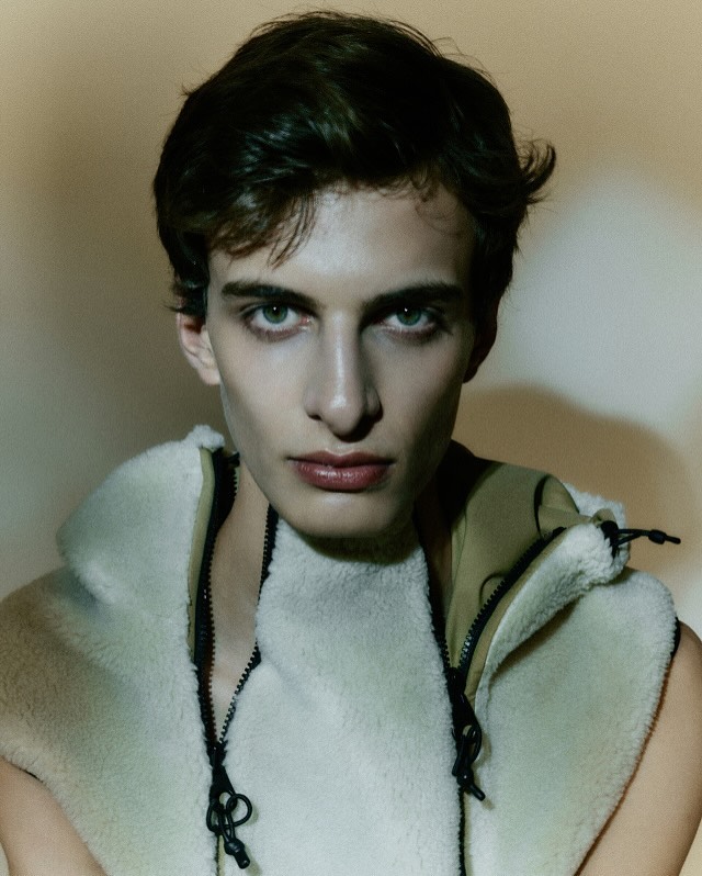Close-up portrait of a young man with dark hair and light eyes, wearing a fleece vest. The soft lighting highlights his features.