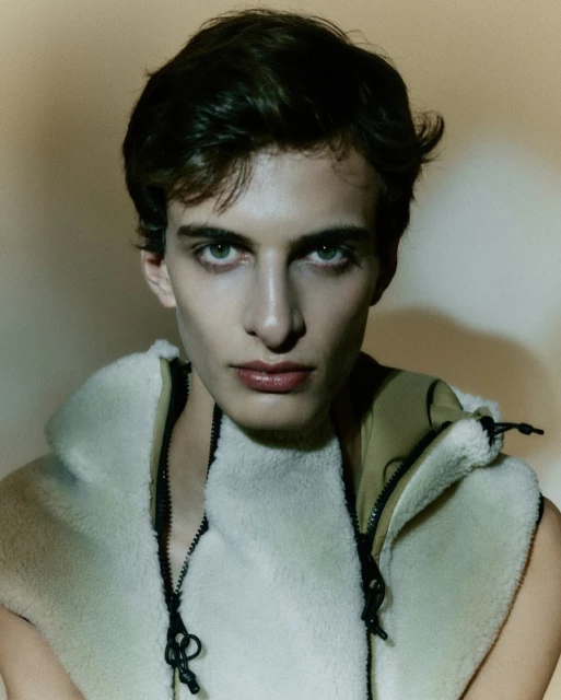 Close-up portrait of a young man with dark hair and light eyes, wearing a fleece vest. The soft lighting highlights his features.