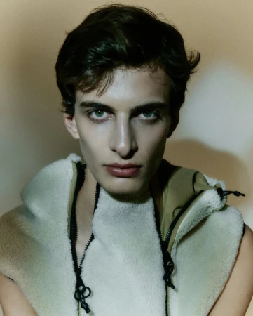 Close-up portrait of a young man with dark hair and light eyes, wearing a fleece vest. The soft lighting highlights his features.