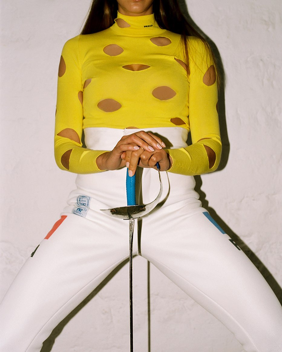 Fashion shot of a woman posing in white fencing pants and a yellow shirt with cutouts, holding a fencing foil.