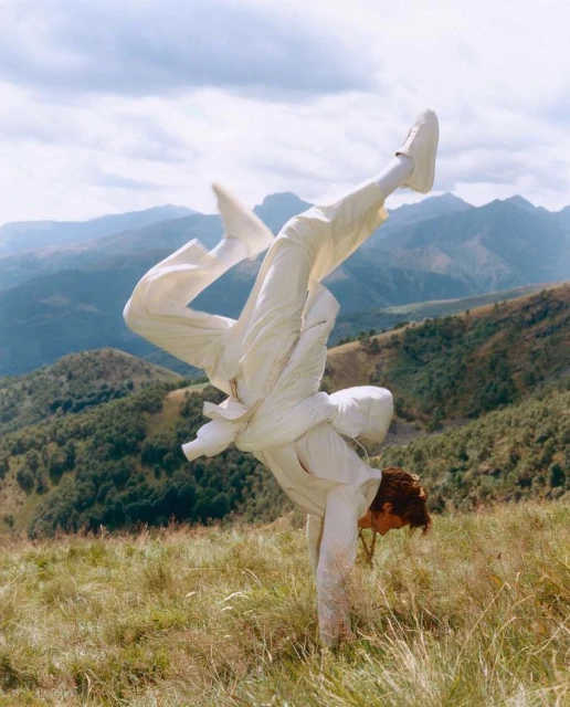 An adult in white clothing performs a handstand in a grassy field with a mountain range and cloudy sky in the background.