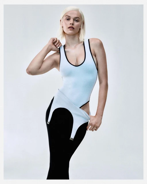 A fair-skinned adult female model with short platinum blonde hair poses in a light blue bodysuit and a black skirt against a gray backdrop.