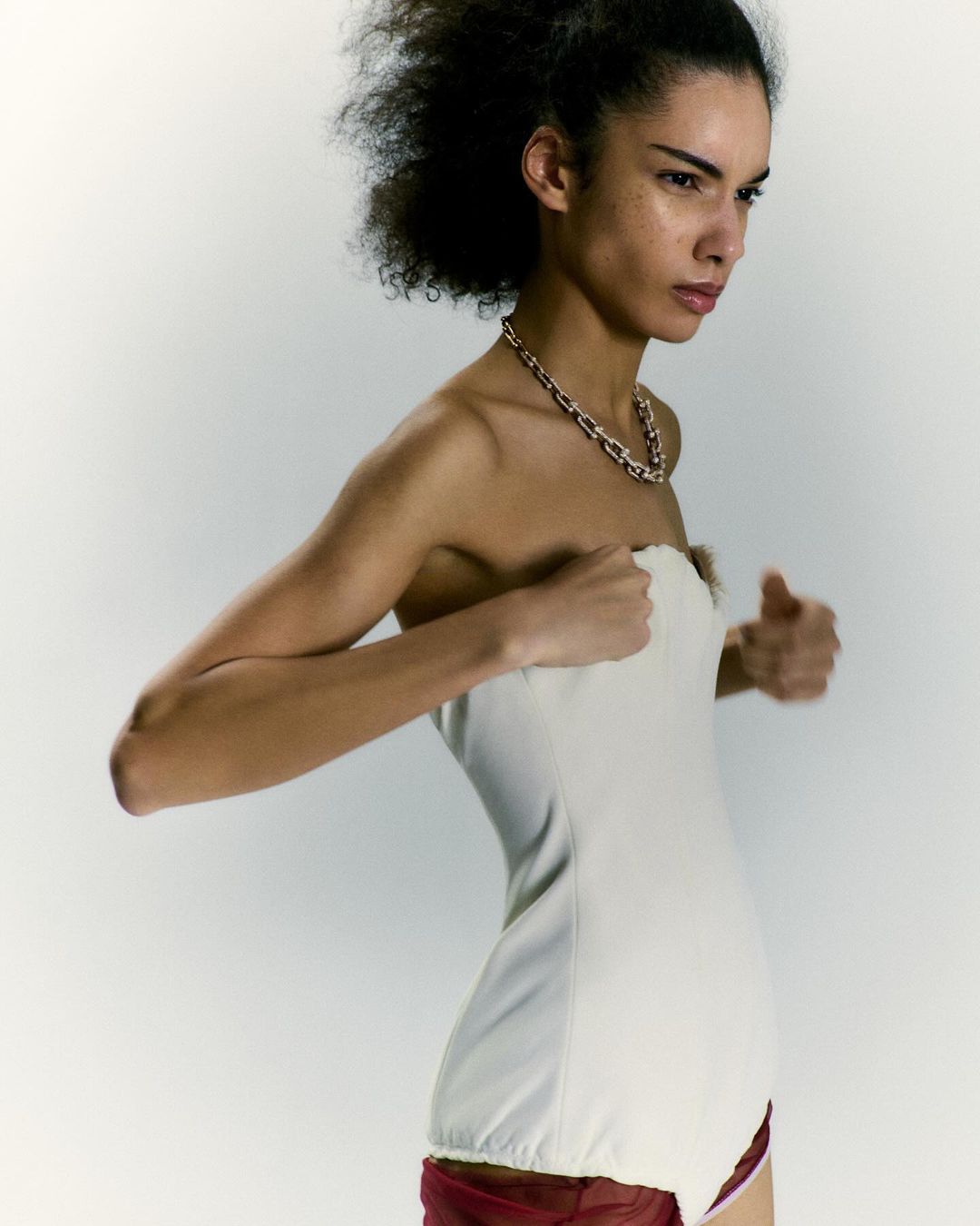 Model with dark curly hair wearing a white strapless top and a gold chain necklace against a light grey background.