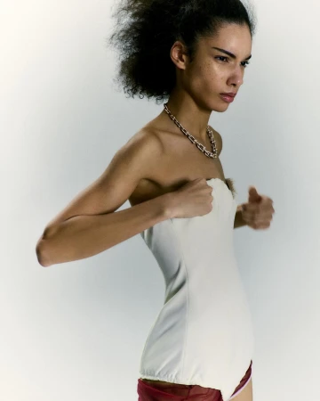 Model with dark curly hair wearing a white strapless top and a gold chain necklace against a light grey background.