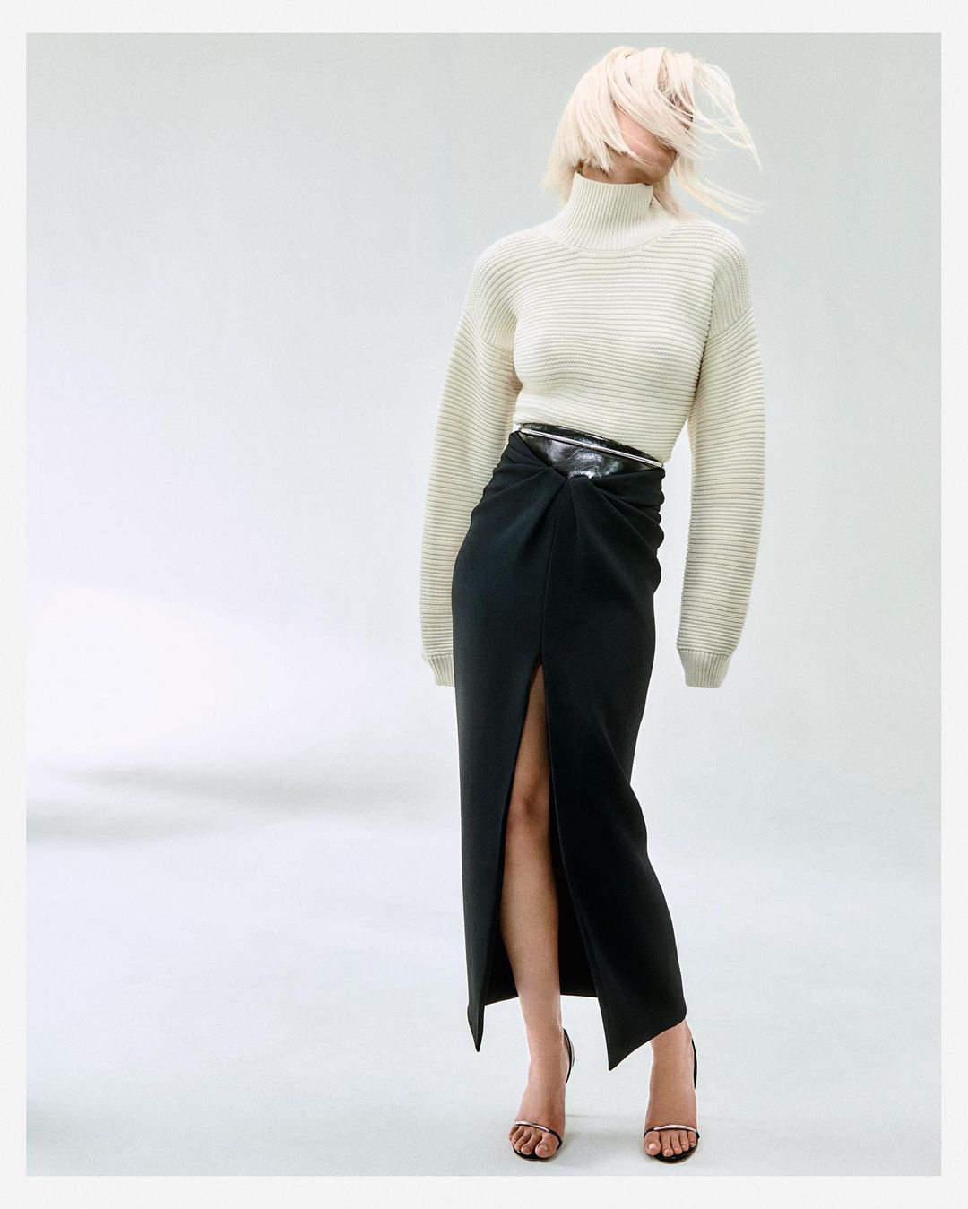 Model with blonde hair whipping across face, wearing ribbed white turtleneck and black slit skirt by Matthieu Delbreuve