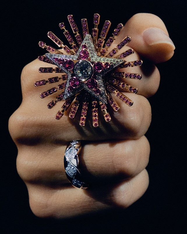 Hand displaying two rings; one star-shaped cocktail ring with diamonds and red gemstones, and one with a braided design. Dark background.