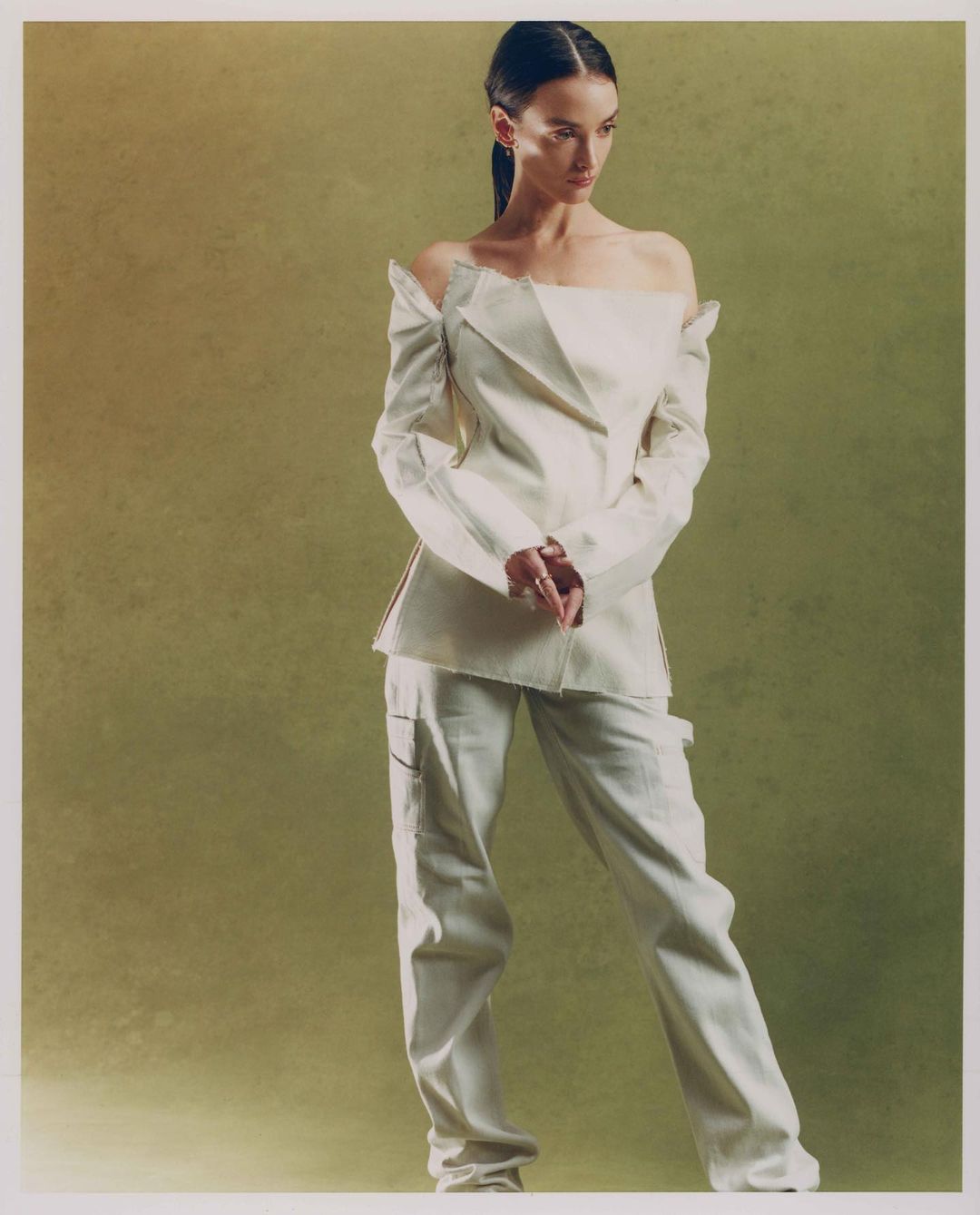 A woman in an off-shoulder jacket and pants poses against a textured green backdrop in a studio setting with soft, diffused lighting.