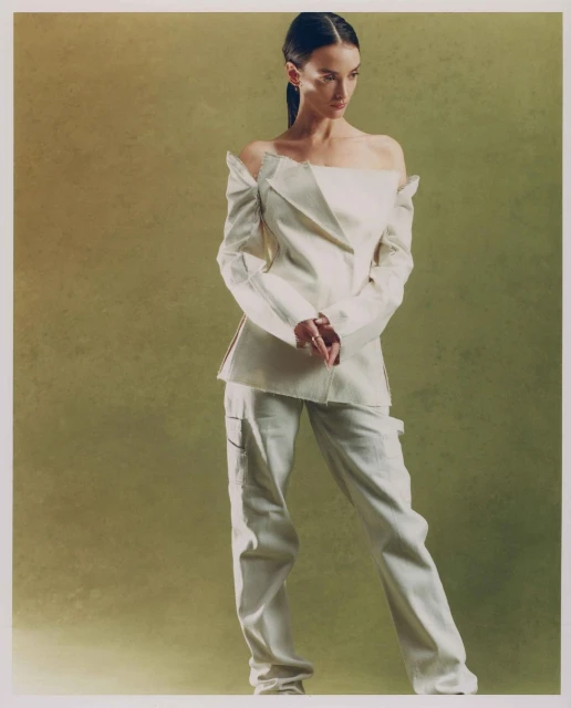 A woman in an off-shoulder jacket and pants poses against a textured green backdrop in a studio setting with soft, diffused lighting.