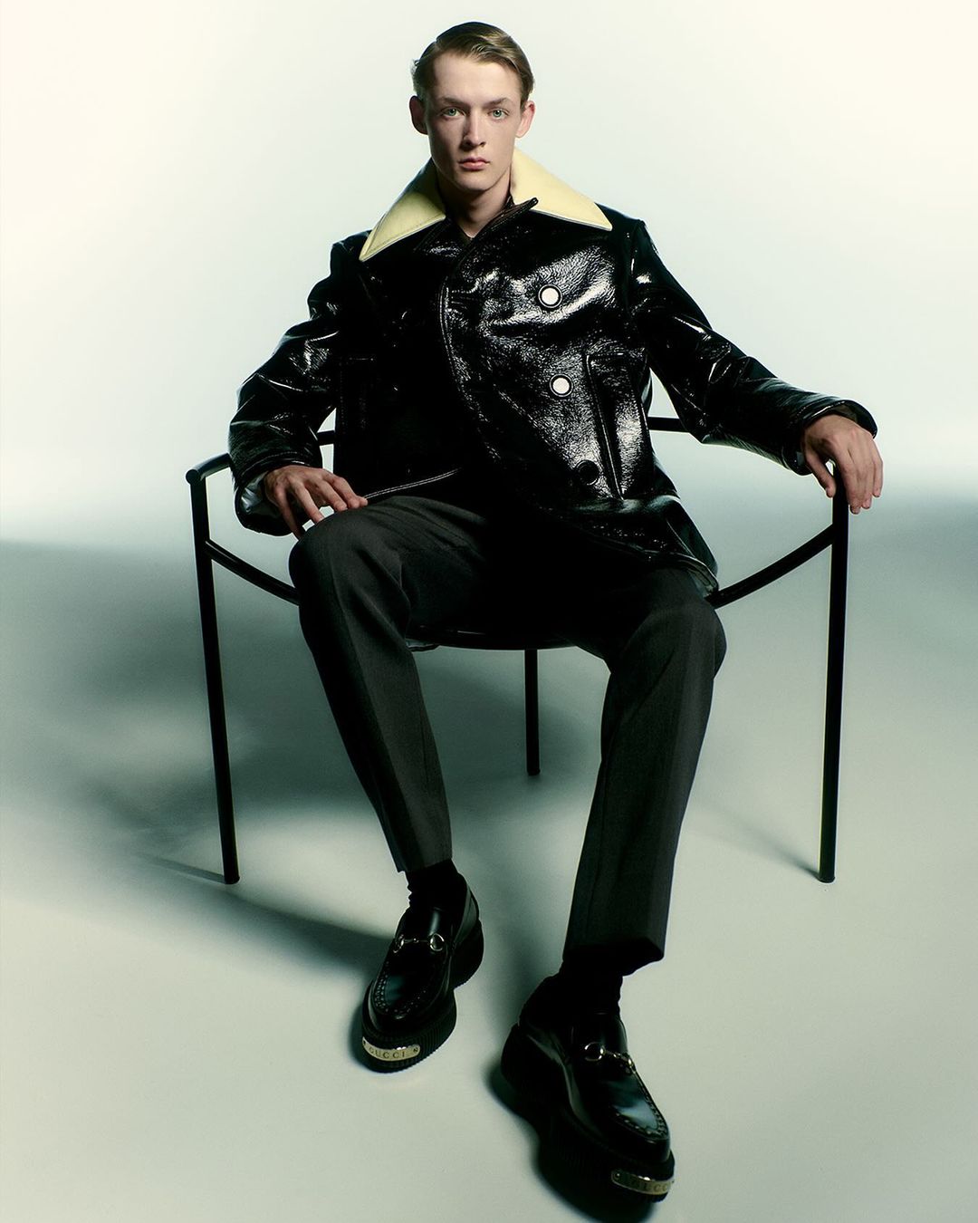 Fashion shot of a young adult man in a black, glossy jacket with a shearling collar, grey pants and dress shoes sitting in a chair.