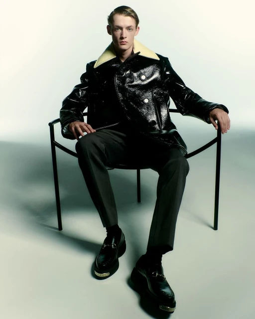 Fashion shot of a young adult man in a black, glossy jacket with a shearling collar, grey pants and dress shoes sitting in a chair.