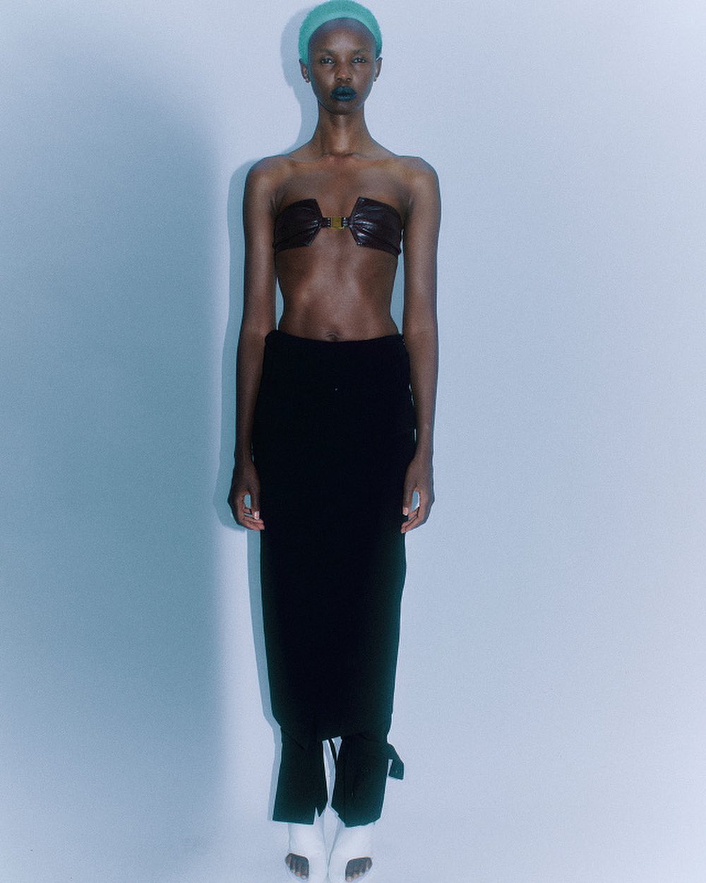 Model with a green cap wearing a dark strapless top and black skirt against a pale wall.