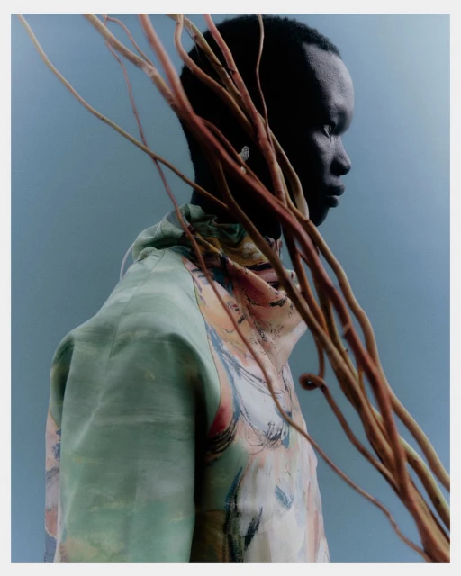 Profile view of a Black adult male wearing a colorful patterned shirt, styled with bare branches against a blue-grey background.