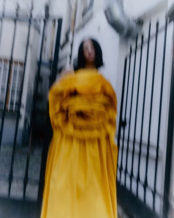Motion blurred portrait of a person in a voluminous bright yellow dress standing outdoors near iron gates in Paris by Matthieu Delbreuve