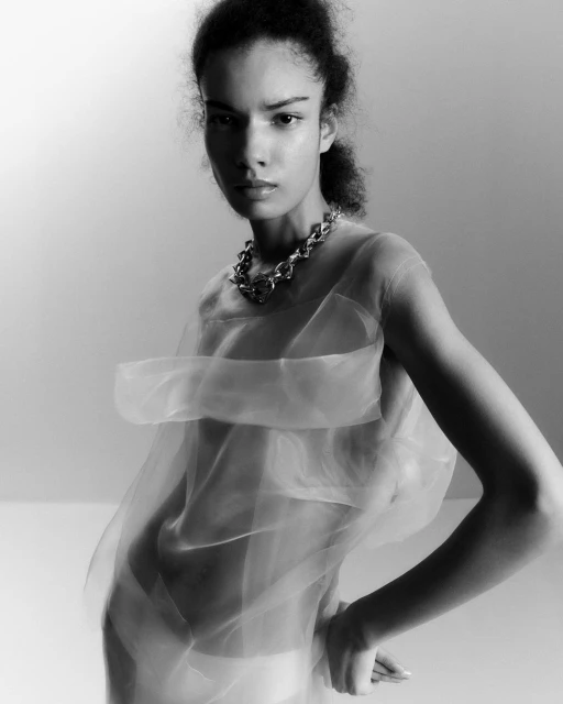 Black and white portrait of an adult woman in a sheer top and chainlink necklace with her hands on her hips in a studio setting.