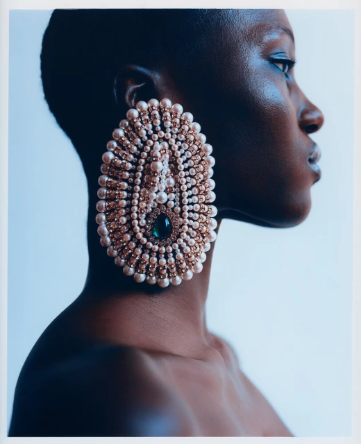 Profile portrait of Black woman wearing a large, pearlescent statement earring with a green stone against a light blue background.