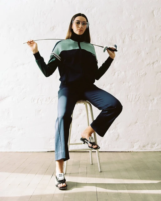Young woman in a black and mint green track suit poses sitting on a metal stool holding a fencing foil in a brightly lit indoor studio.