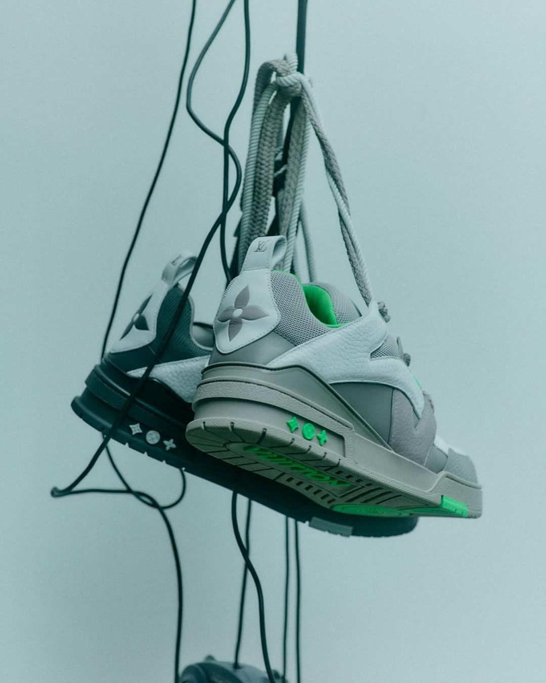 Grey sneakers hanging by laces from vertical wires against a neutral grey studio background