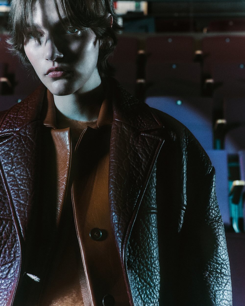 Portrait of a young adult with dark hair wearing a brown leather jacket and brown top, posing indoors under cool blue lighting.