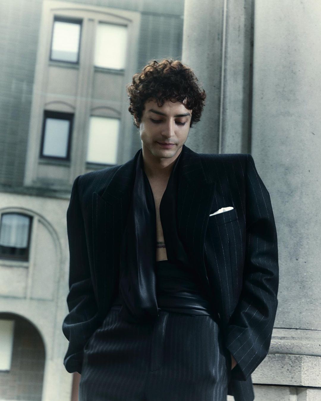 Portrait of a young adult man in a pinstripe suit stands outdoors against a building, looking down with neutral facial expression.