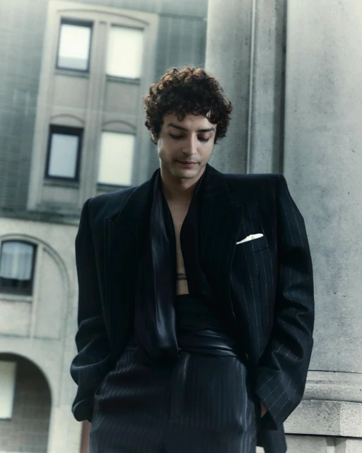 Portrait of a young adult man in a pinstripe suit stands outdoors against a building, looking down with neutral facial expression.