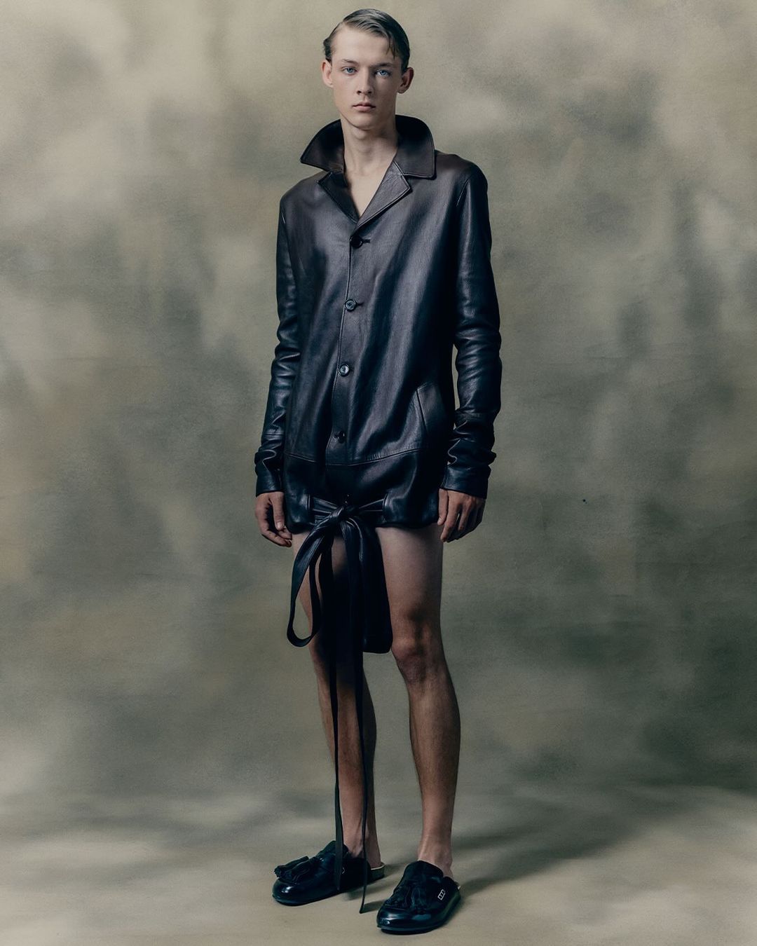 Fashion portrait of a young adult male wearing a dark leather-like shirt and black slide sandals against a neutral background. 