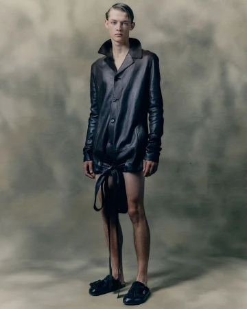 Fashion portrait of a young adult male wearing a dark leather-like shirt and black slide sandals against a neutral background. 