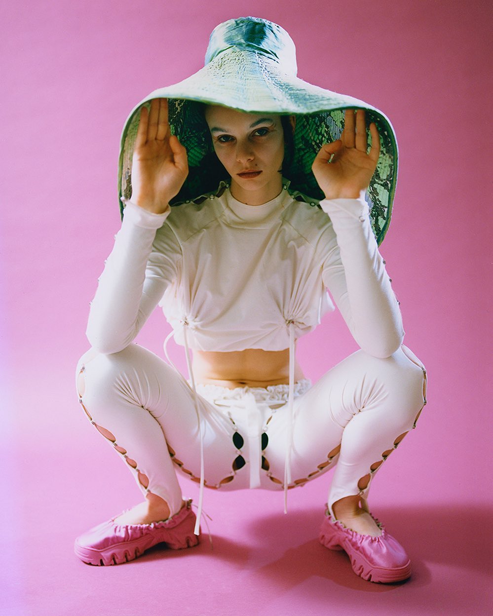 A stylish woman in white squatting with a unique hat and shoes on a pink background in a fashion photography studio setting.