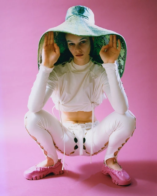 A stylish woman in white squatting with a unique hat and shoes on a pink background in a fashion photography studio setting.