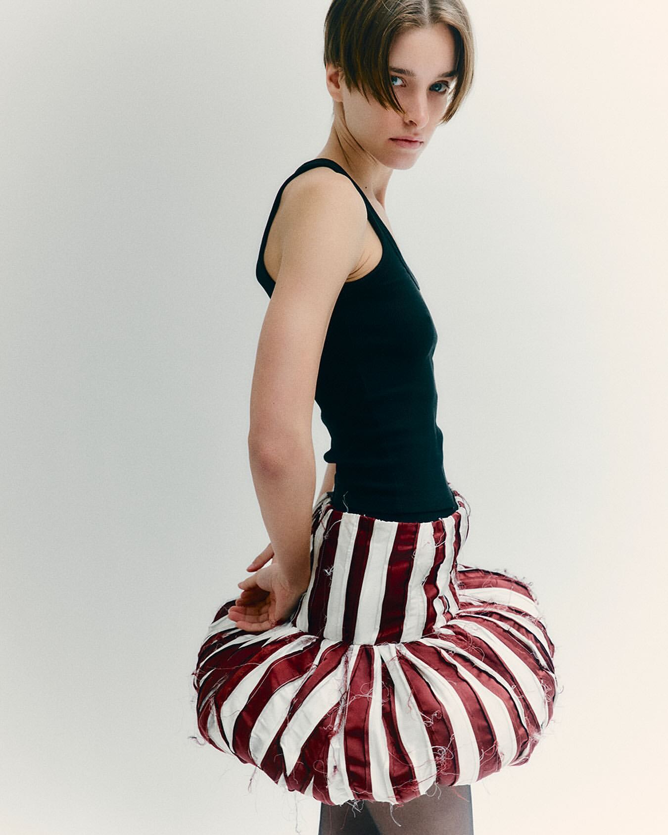 Fashion shot of a woman wearing a black top and sculptural red and white striped skirt, looking at the camera in a studio setting.