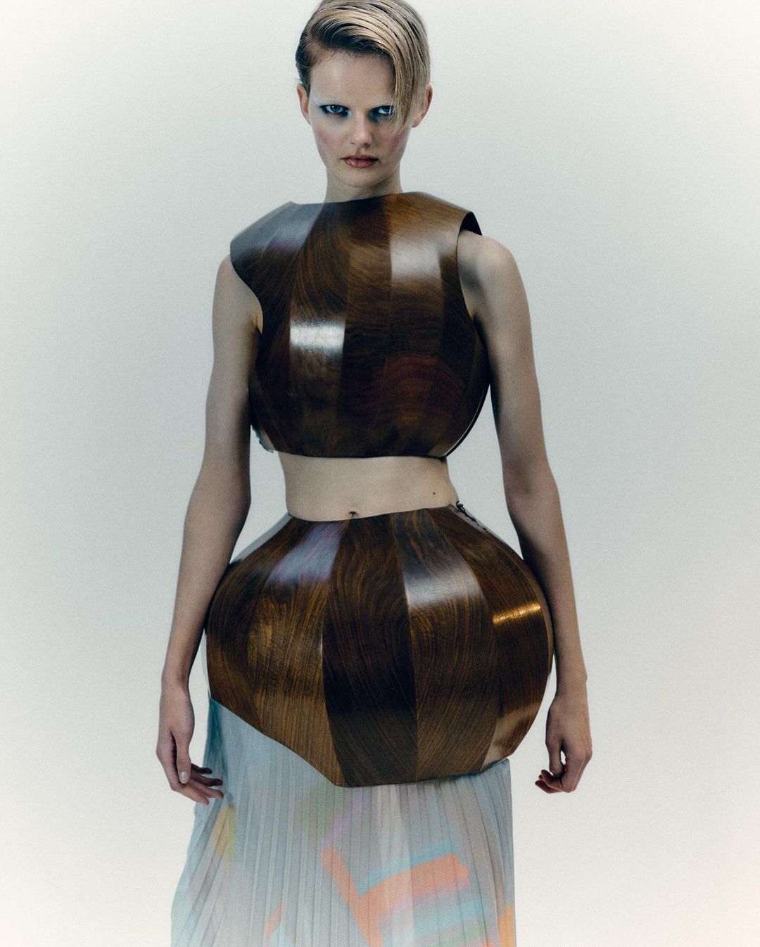 A model poses in a futuristic, geometrically shaped wooden crop top and skirt with a pleated pastel skirt in a studio setting.
